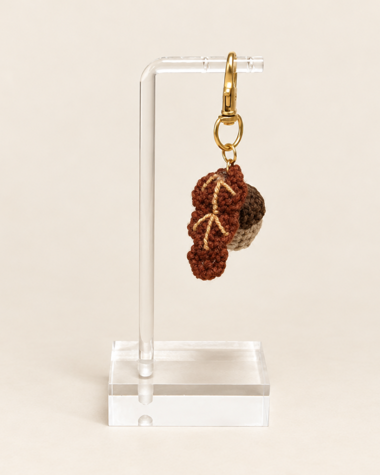 Ecru acorn with a dark brown cap beside a brown oak leaf, attached to a gold-tone clasp on a clear stand.