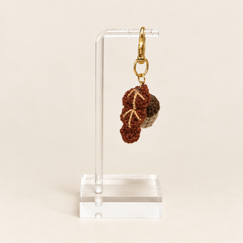 Ecru acorn with a dark brown cap beside a brown oak leaf, attached to a gold-tone clasp on a clear stand.